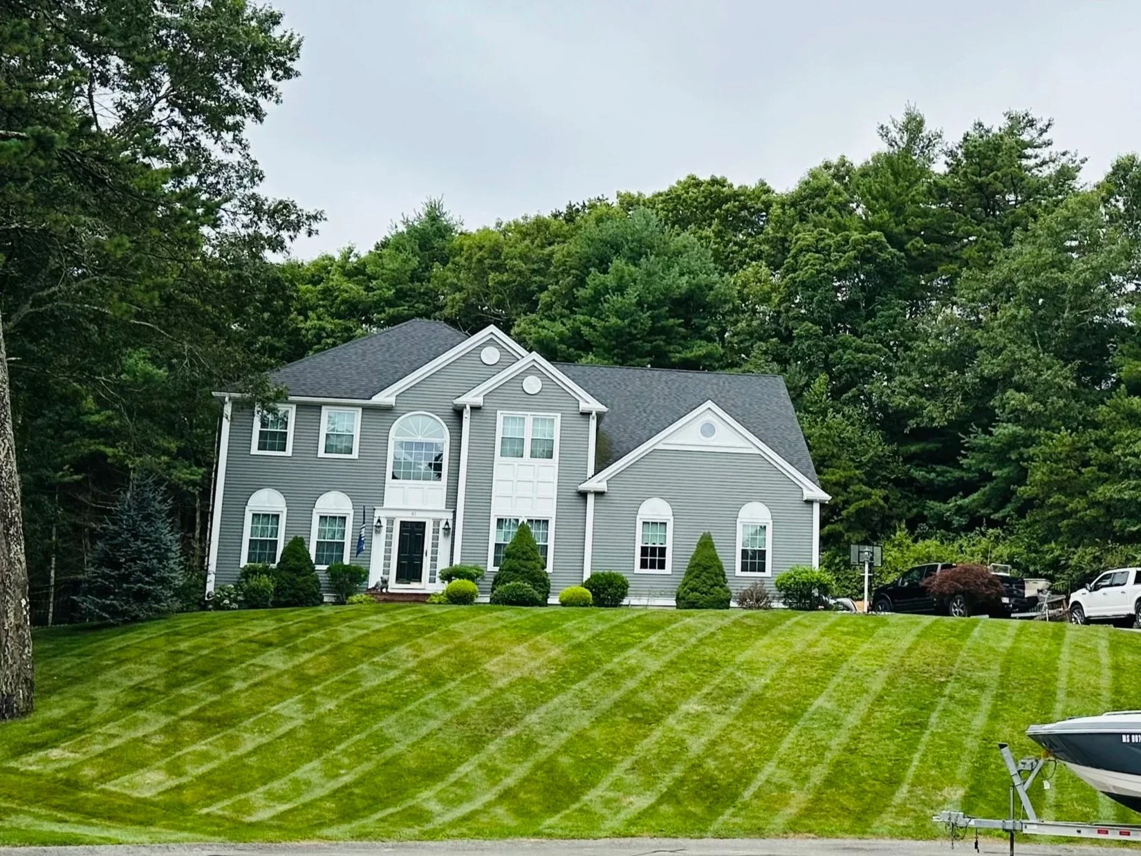 A large gray two-story house with a well-maintained, striped lawn and surrounded by trees. There are multiple windows, bushes, and a driveway with cars parked. A boat is partially visible in the foreground.