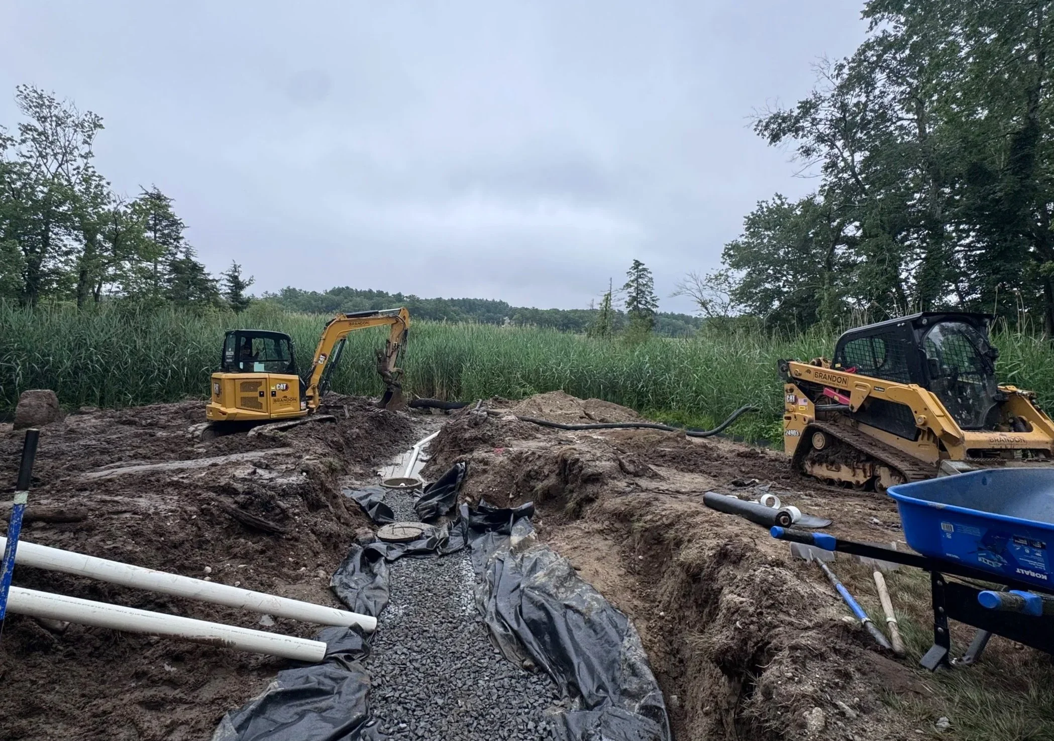 Construction site with two small excavators working on underground piping in a rural area with tall grass and trees in the background, overcast sky.