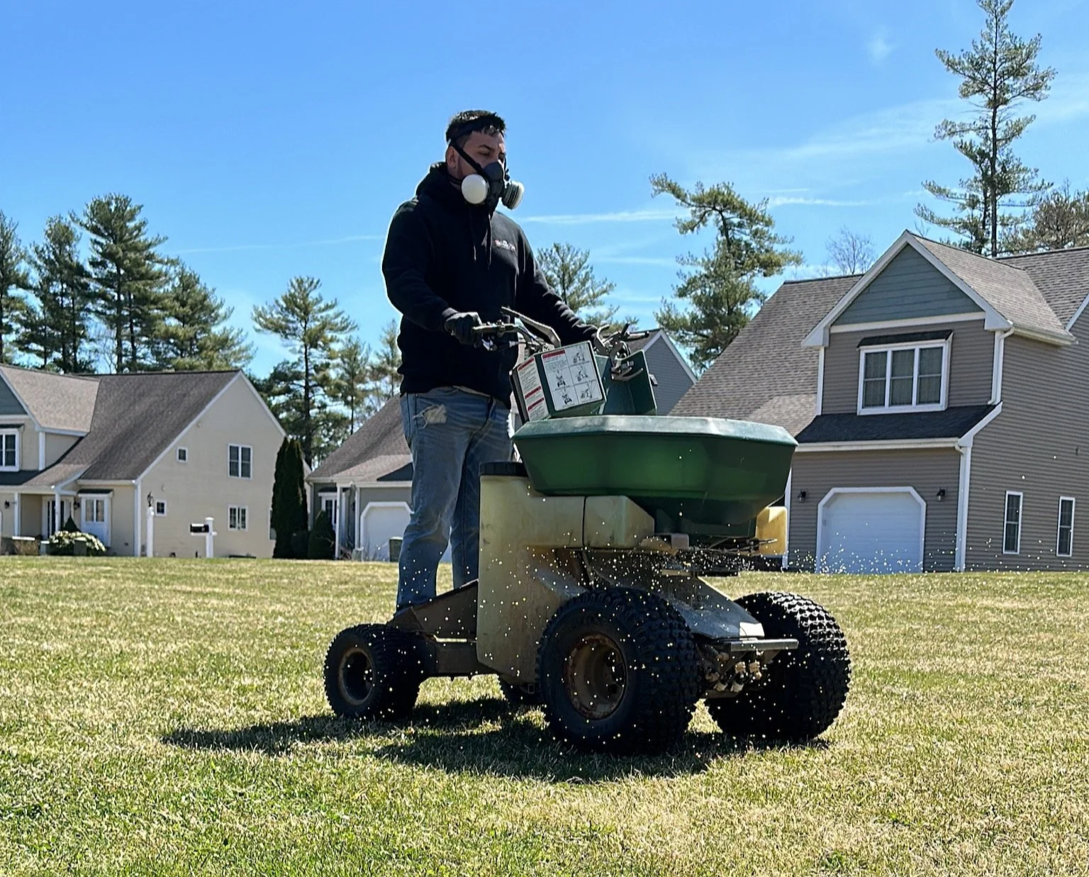 Person wearing a face mask, black hoodie, and gloves operating a small green and black lawn sprayer on a grassy field in a neighborhood with houses and trees.