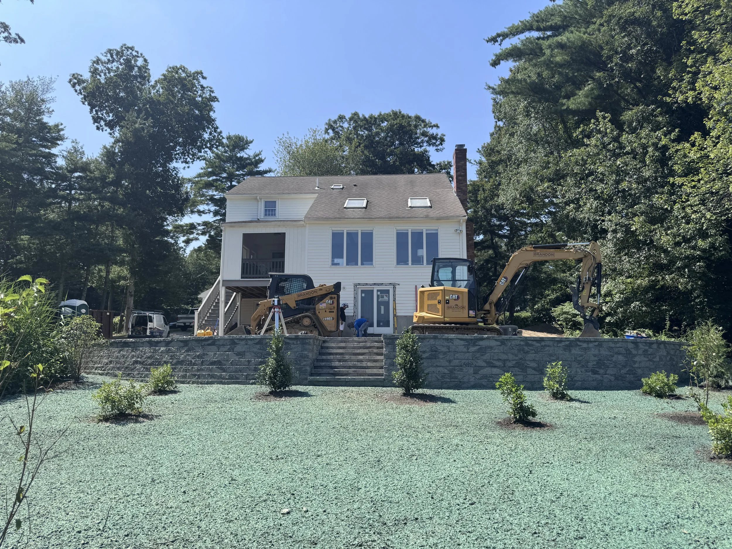 Construction workers and equipment on a raised patio of a two-story white house with large windows, surrounded by trees and a gravel yard.