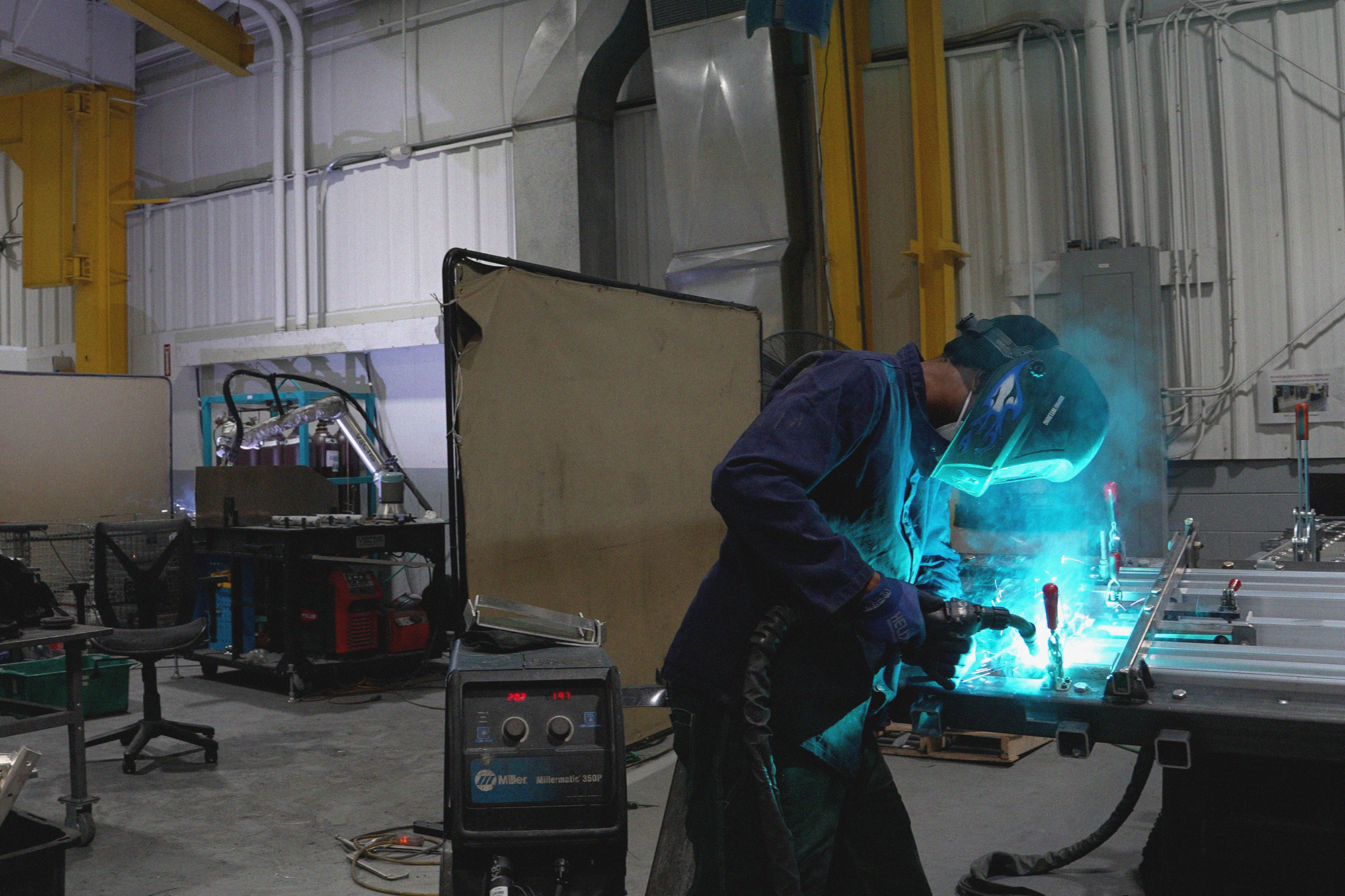 Welder working at an efficient Caltech Manufacturing workstation designed for quick part changeover, with a Vectis cobot welding system at a nearby fixture table.