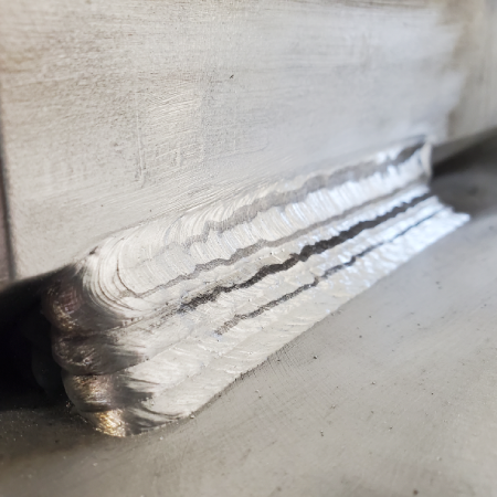 Close-up of a multi-pass aluminum fillet weld joining a vertical member to a base plate, showing distinct weld passes and complete joint coverage.