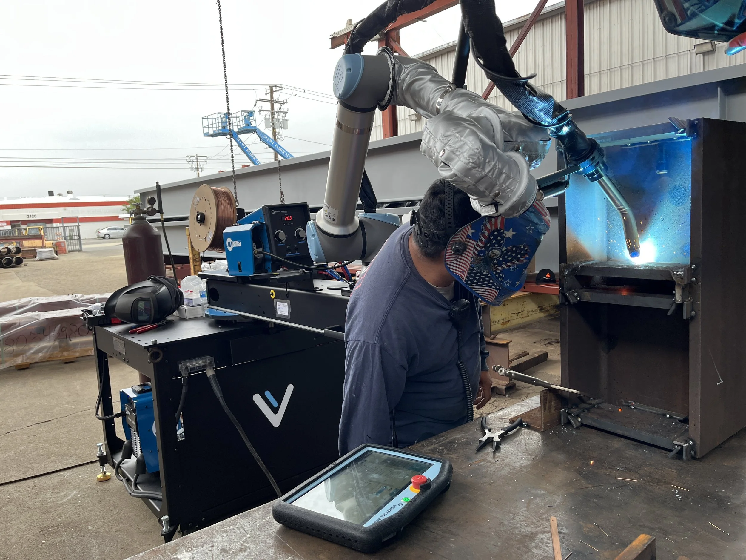 Technician using a robotic welding arm outdoors, creating a bright blue arc on a metal workpiece, with an automated welding system and tablet control.