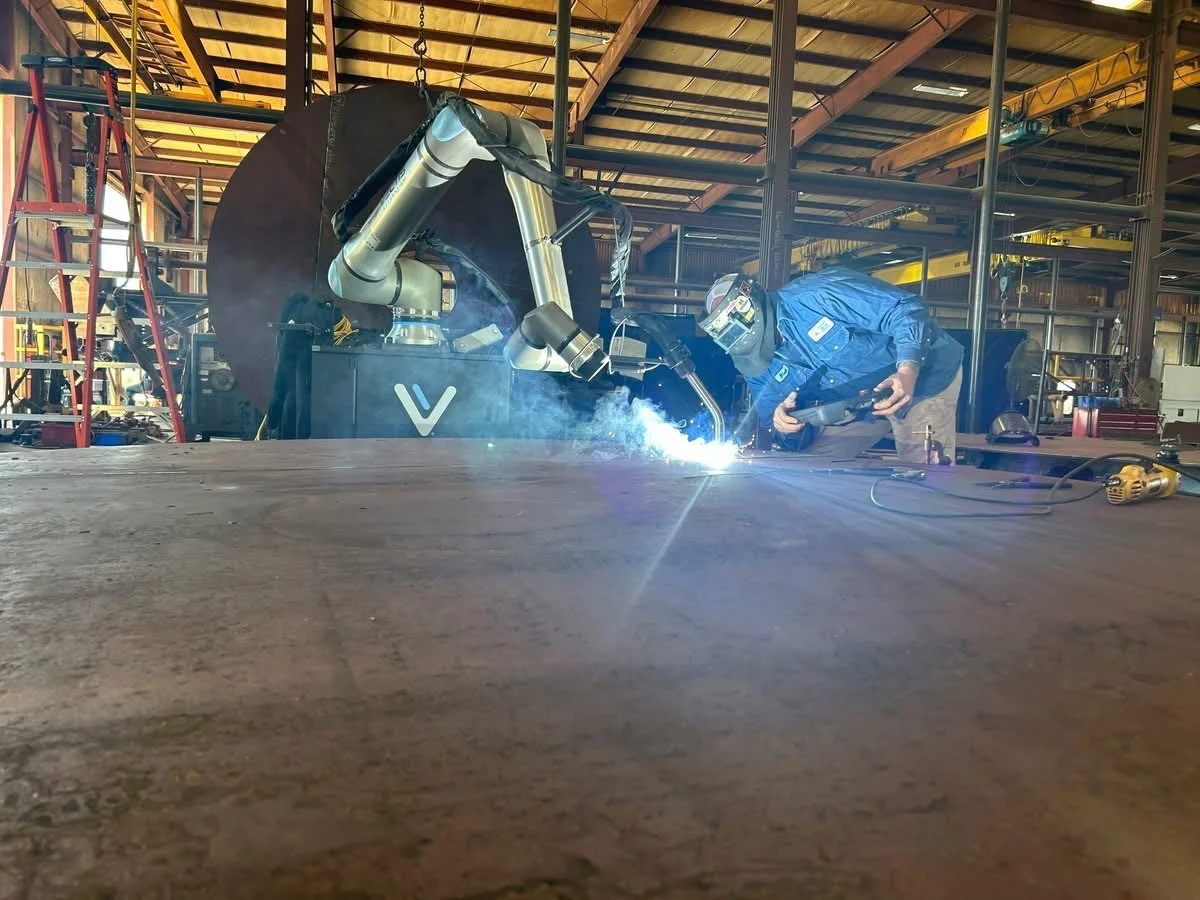 Welder monitors a Vectis cobot welding system as it performs a weld on a large steel fabrication in an industrial shop.