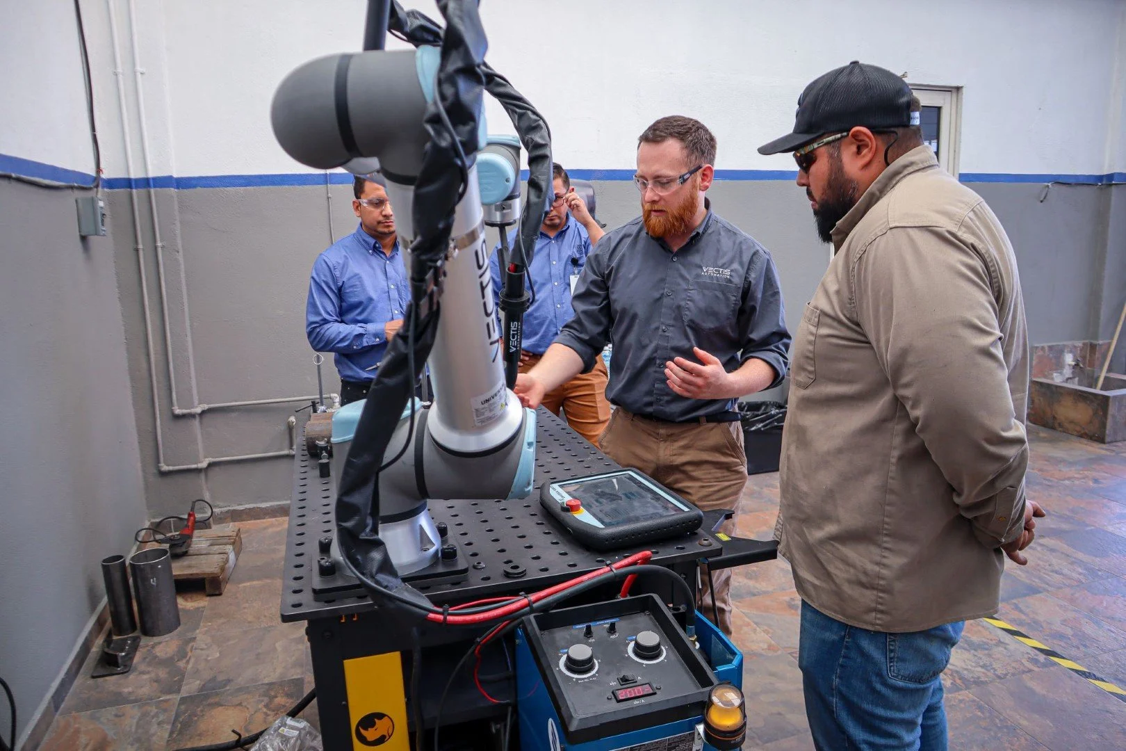 Three professionals observing a robotic arm as one demonstrates controls, highlighting hands‑on industrial automation training in a workshop.