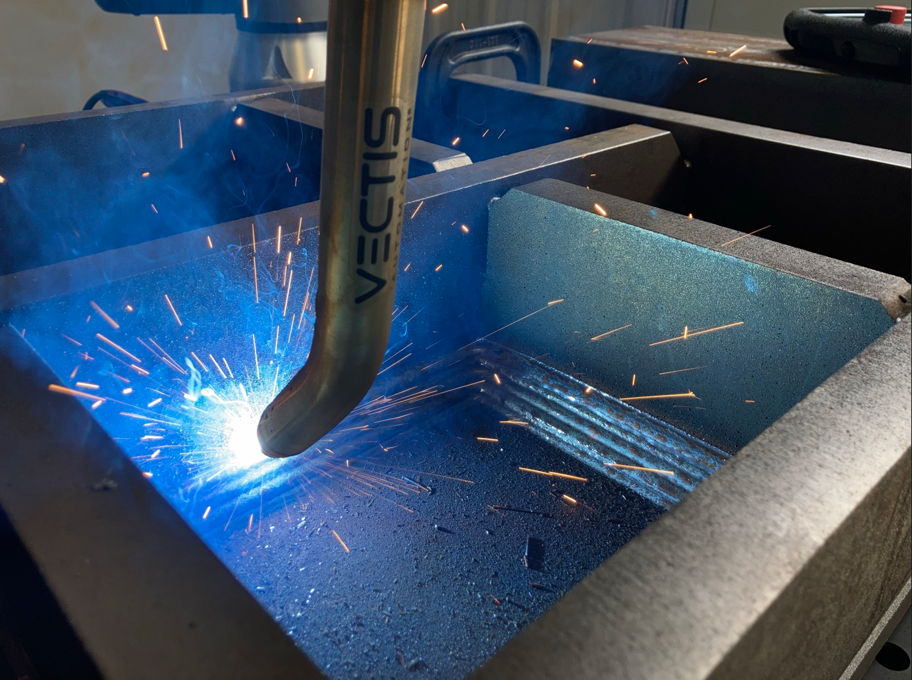 Robotic welding torch creating a bright blue arc and sparks while joining a metal seam inside a steel fixture, showcasing automated industrial welding.