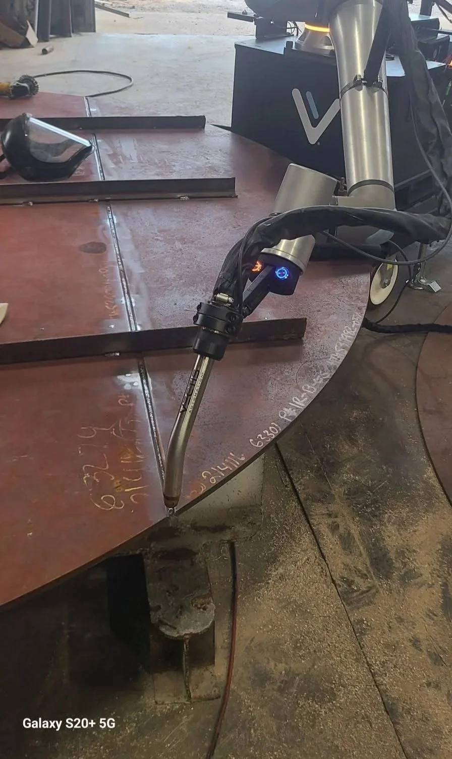 Close-up of a Vectis cobot welding torch positioned over a curved steel plate with long weld seams in a fabrication shop.