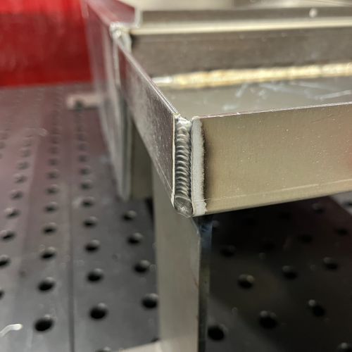 Close-up of an outside-corner fillet weld on a fabricated aluminum box assembly, showing a uniform weld bead at the corner joint.