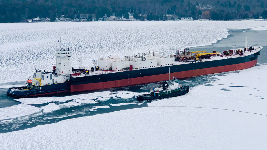 Large vessel escorted by tugboats through an ice-covered waterway.