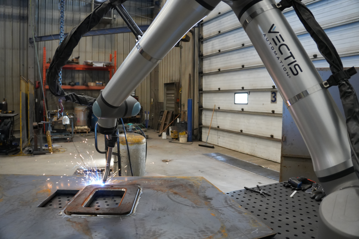 Cobot welding system performing MIG welding on a steel plate with visible sparks in a contract manufacturing shop, demonstrating flexible automation for small-batch production and varied weld applications.