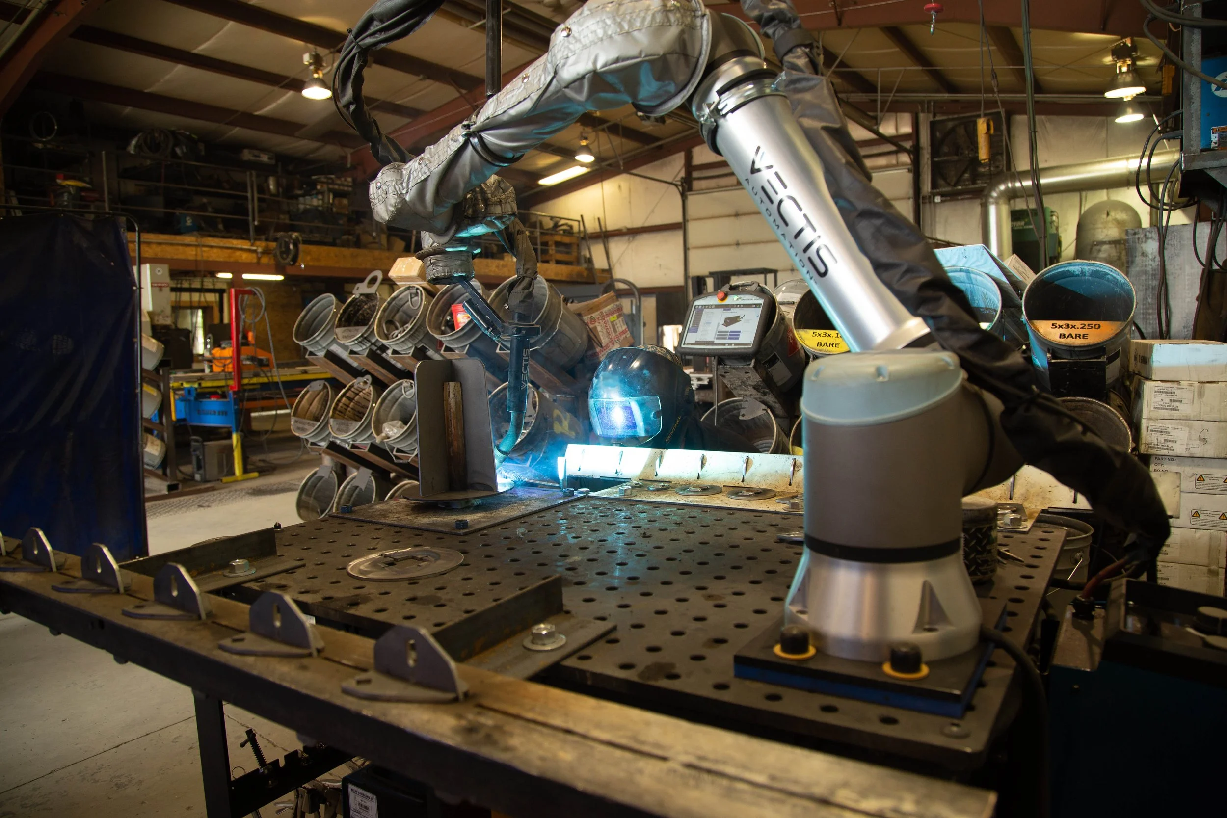 Collaborative robot (cobot) performing arc welding on metal components at a workbench, showing bright welding arc, fixture setup, and automated welding process in an industrial fabrication environment.