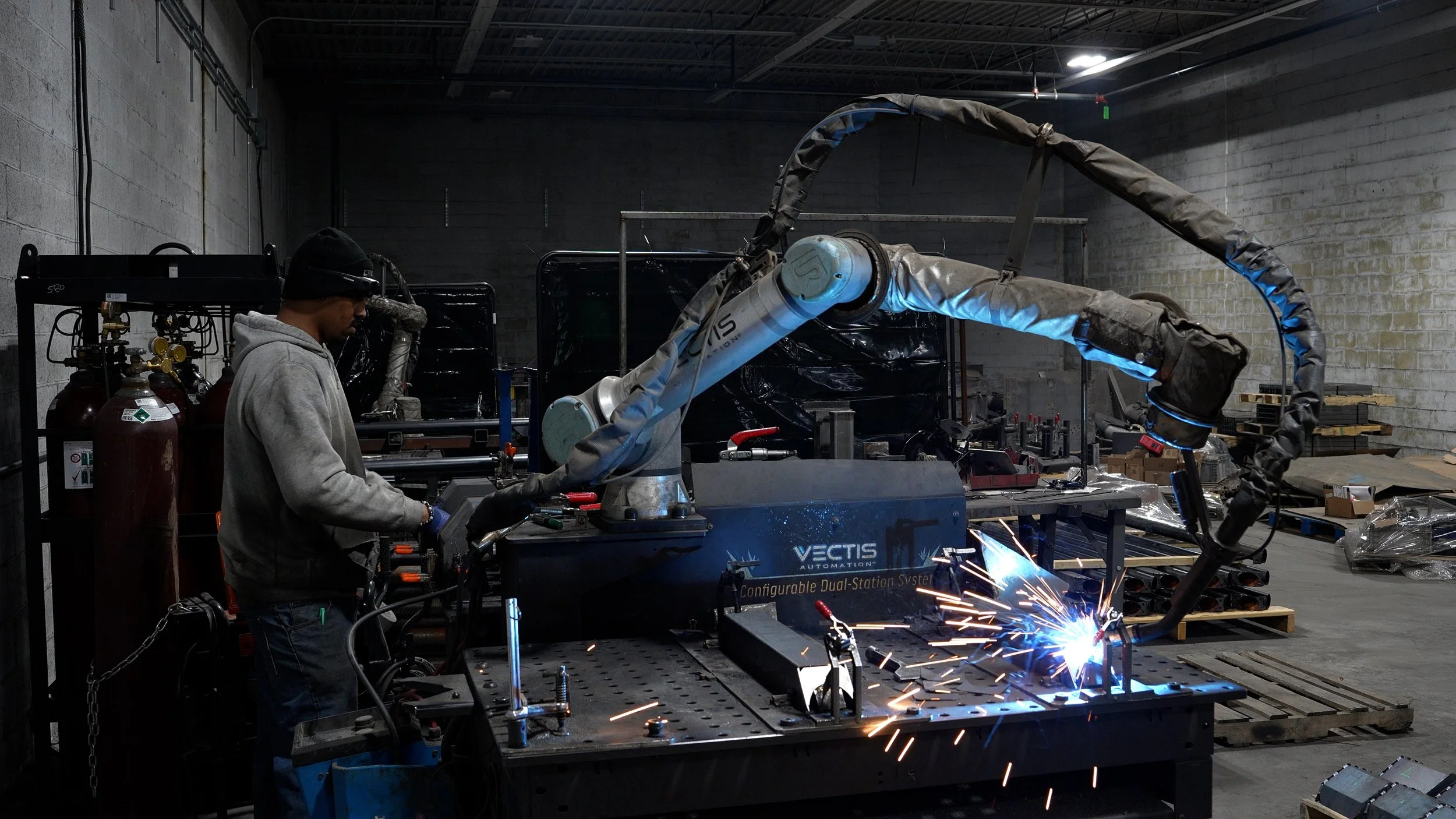 Vectis cobot welding a fixtured metal part at a Caltech Manufacturing dual-station workcell while an operator prepares the next setup.