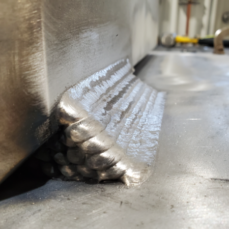 Close-up of a multi-pass aluminum fillet weld joining a vertical component to a base plate, showing a consistent weld bead pattern and complete weld coverage.