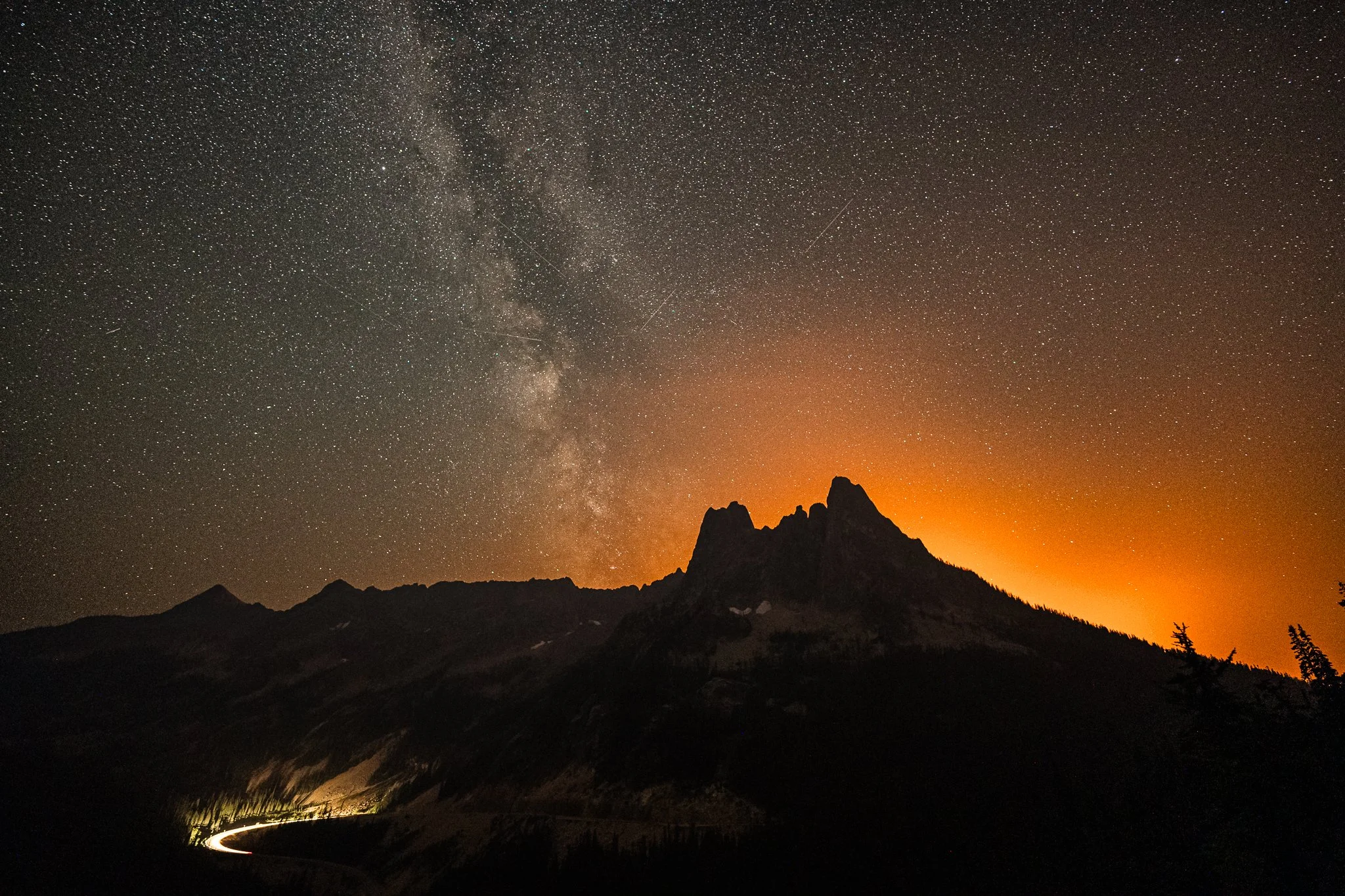 Night sky filled with stars and the Milky Way galaxy above mountain peaks, with a faint orange glow on the horizon and a winding road illuminated at the bottom.