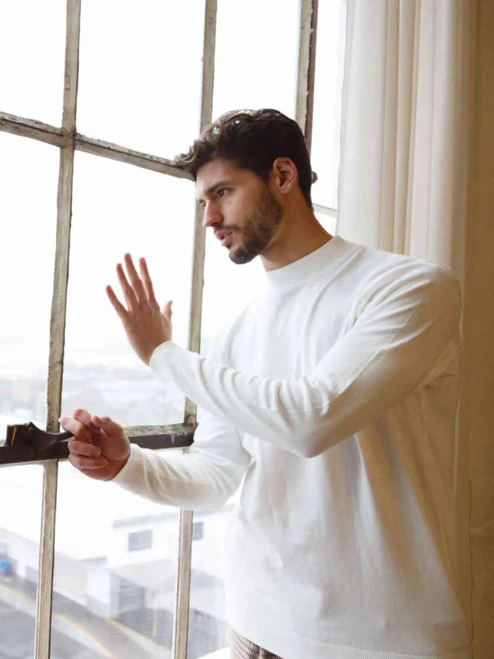 Fashion editorial of a man in a white shirt standing beside a large window
