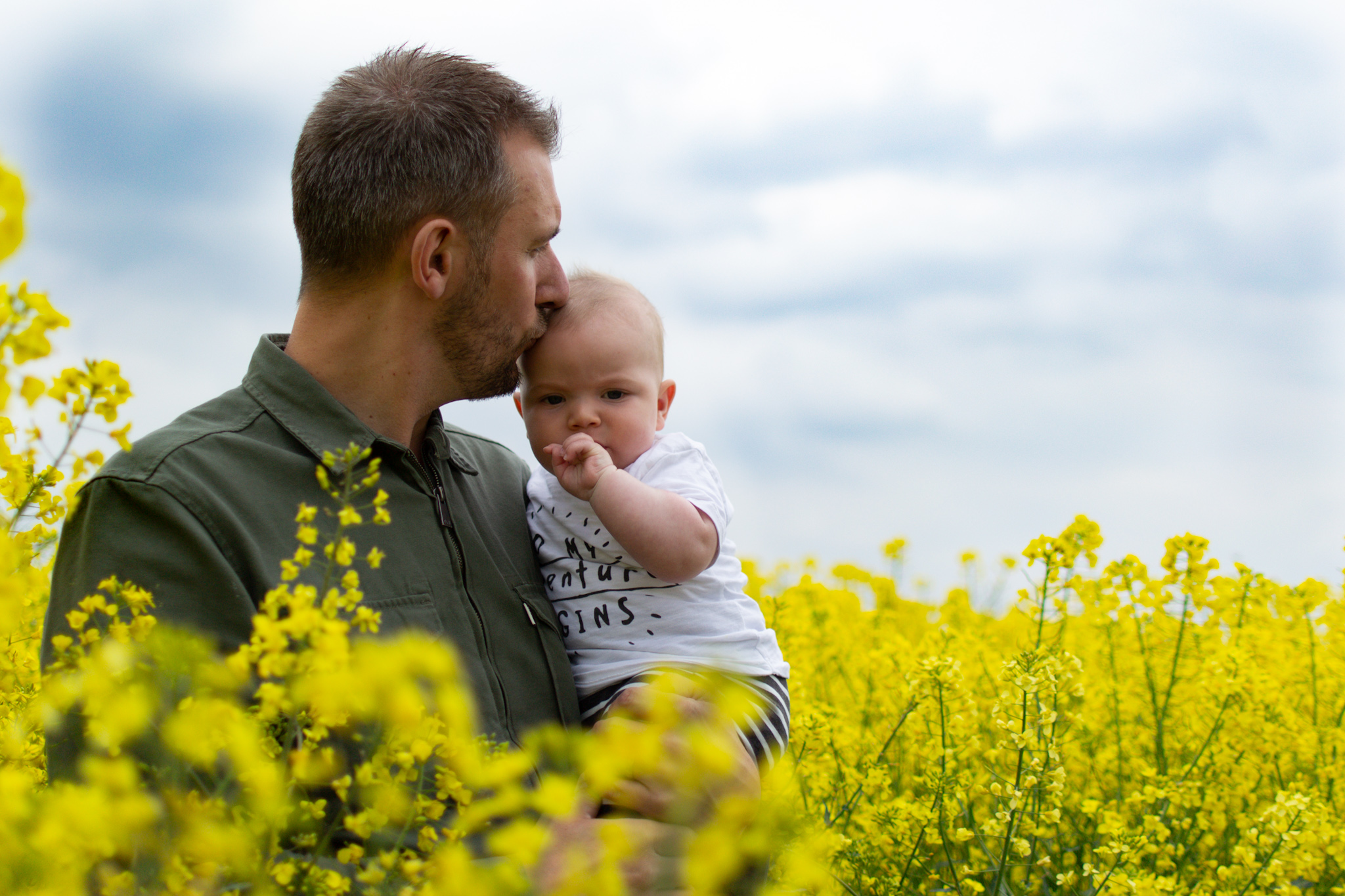  Dad &amp; baby son stood in a field of yellow flowers. Dad is kissing top of sons head. 