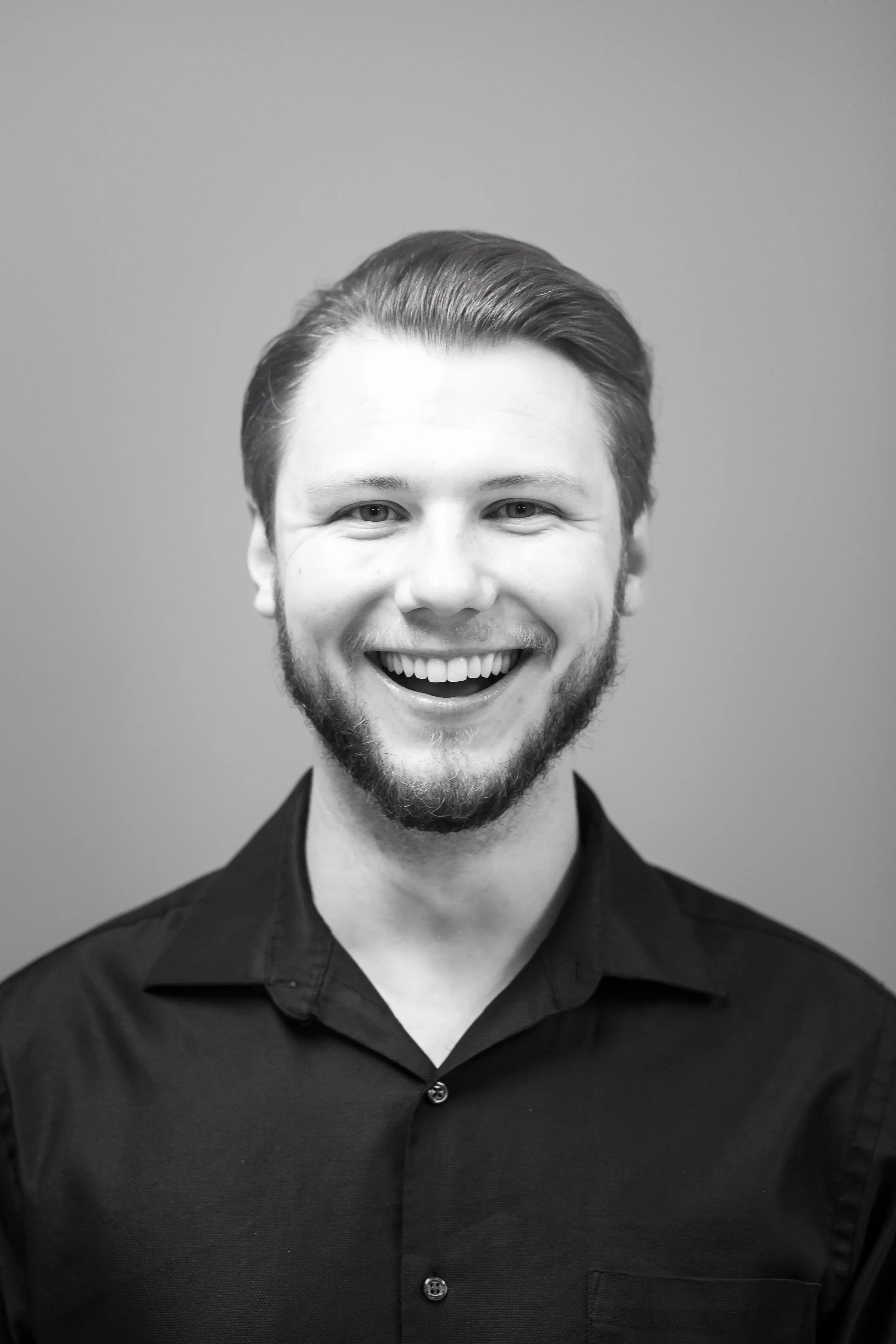 Black and white portrait of a young man with a beard smiling wearing a dark collared shirt against a plain background.