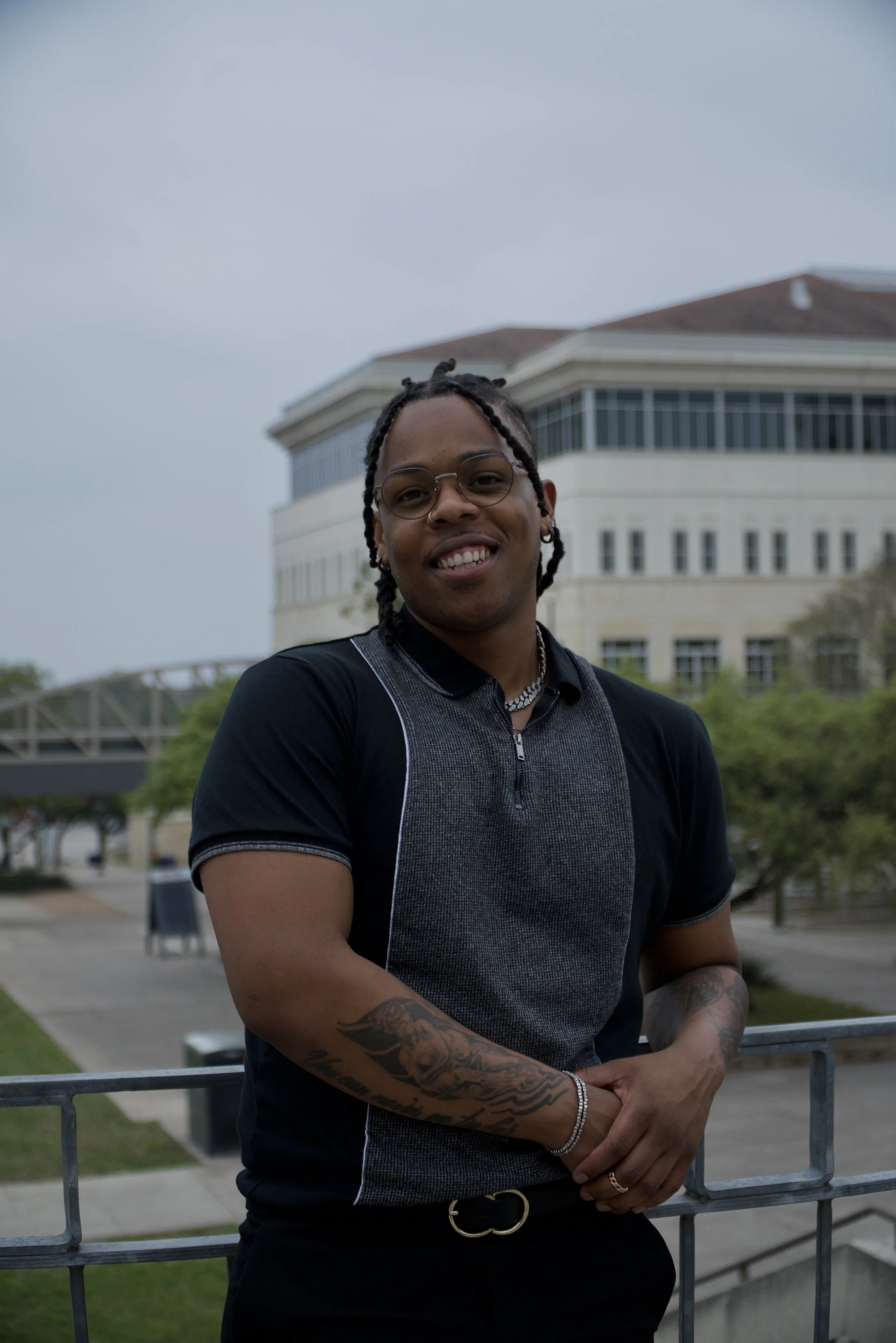 Young man with glasses and tattoos smiling outdoors in front of a modern building and cloudy sky.
