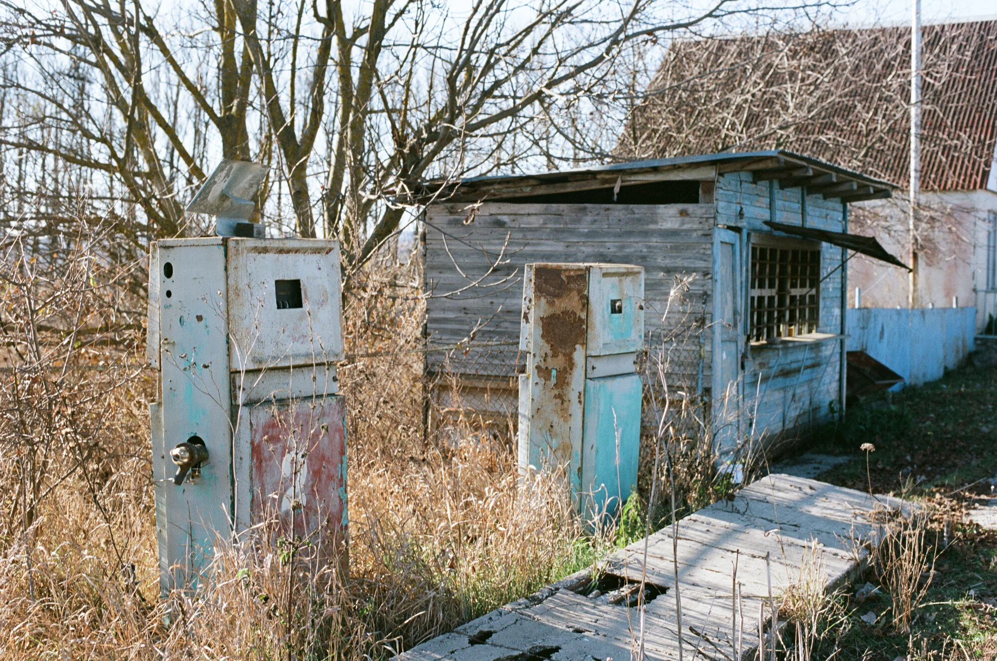 Old, rusted water meters outside an abandoned wooden shack with a broken sidewalk and overgrown grass.