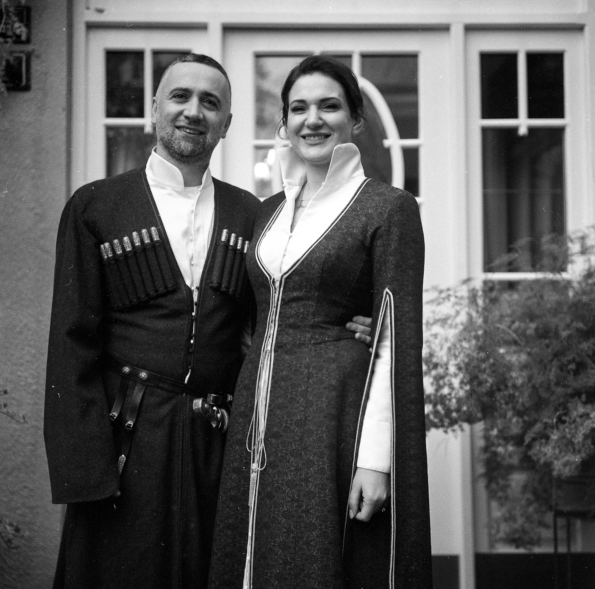 A man and woman dressed in traditional academic or ceremonial robes, standing outdoors in front of a building with large windows, smiling at the camera.