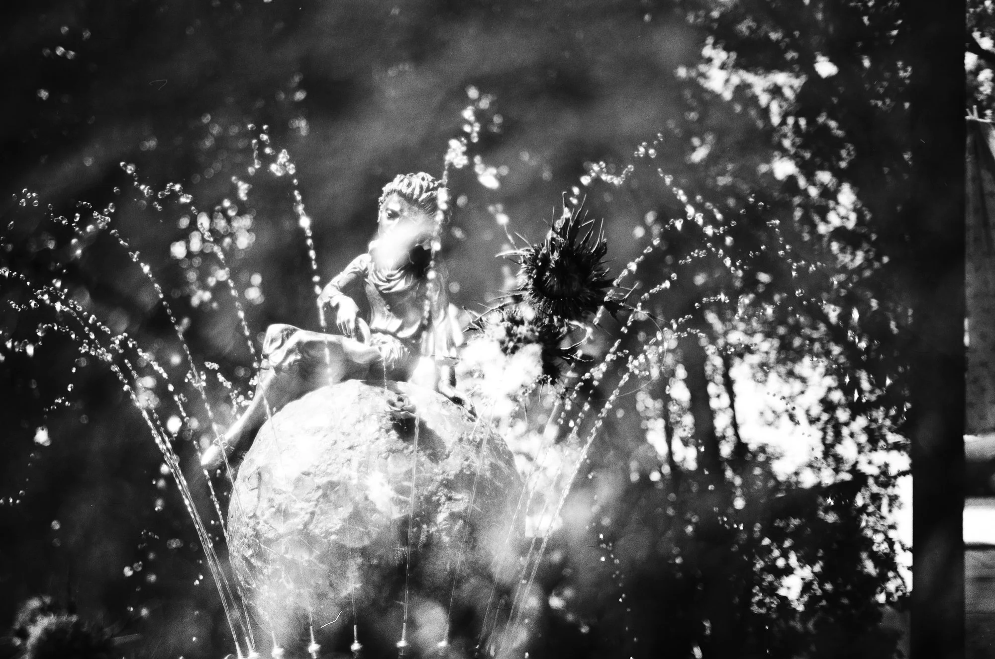 A black and white photograph of a person sitting on a large rock surrounded by a fountain with water spraying in arcs around them. Trees are visible in the background.