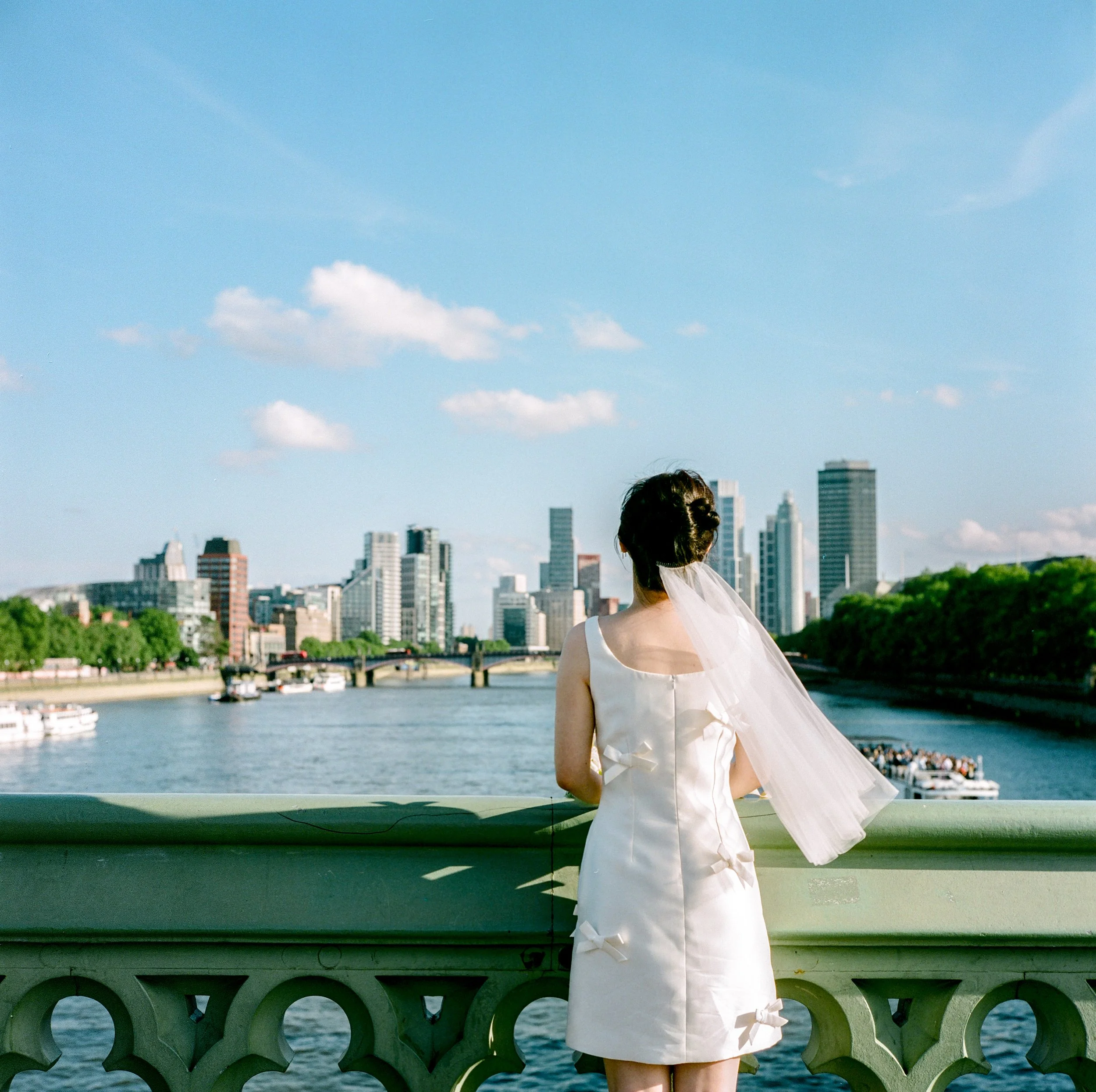 A woman in a white dress standing on a bridge over a river, overlooking a city skyline with tall buildings and boats in the water under a blue sky with scattered clouds.