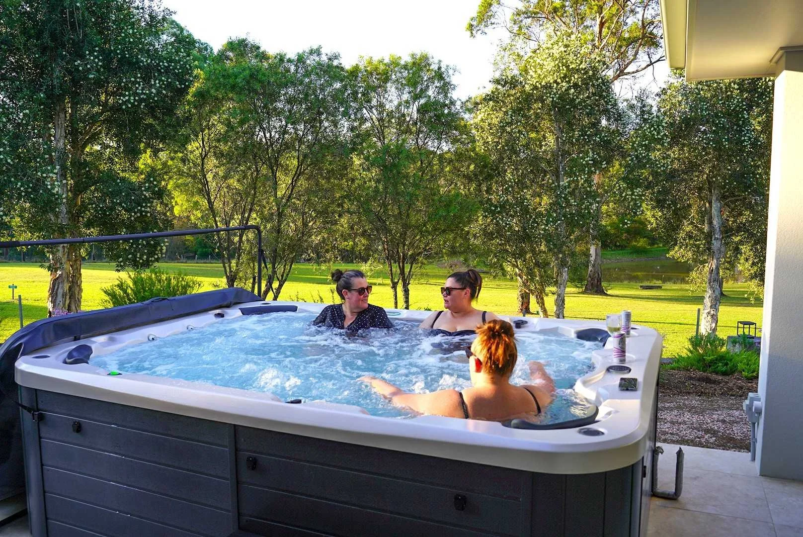 Three women enjoying the outdoor hot tub at Dido’s Place in the Hawkesbury, surrounded by private acreage, lush lawns and trees during golden afternoon light.