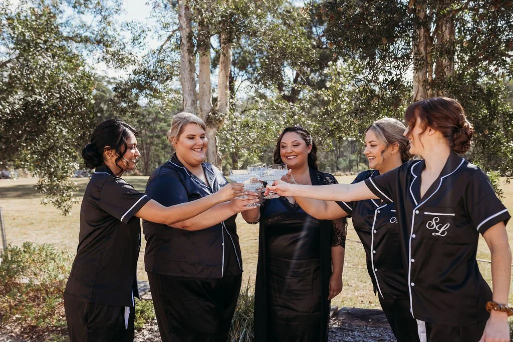 Bride and bridesmaids raising champagne glasses outdoors at Dido’s Place in East Kurrajong, celebrating before their Hawkesbury wedding