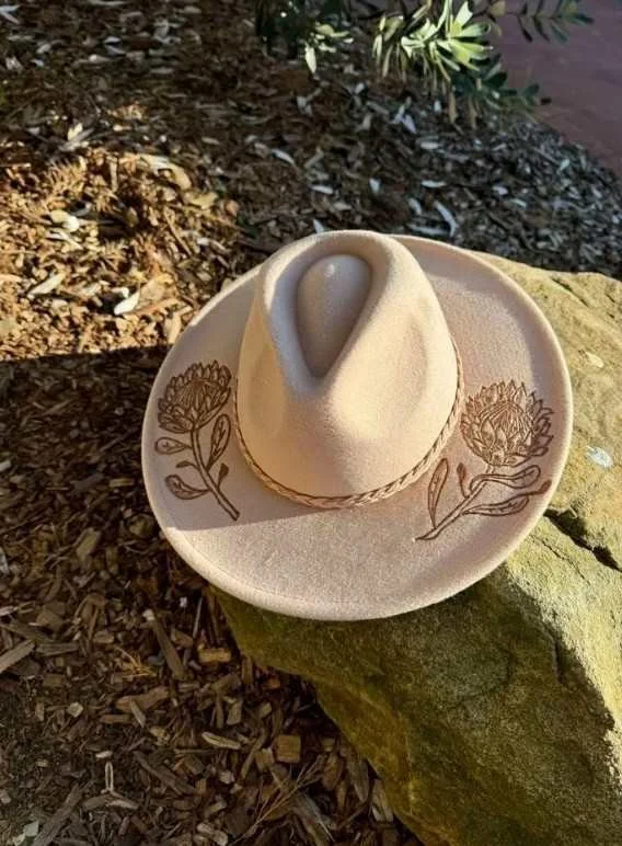 Hand-burned pyrography hat with floral design on a beige felt wide-brim hat, showcasing detailed linework and shading in a creative workshop setting.