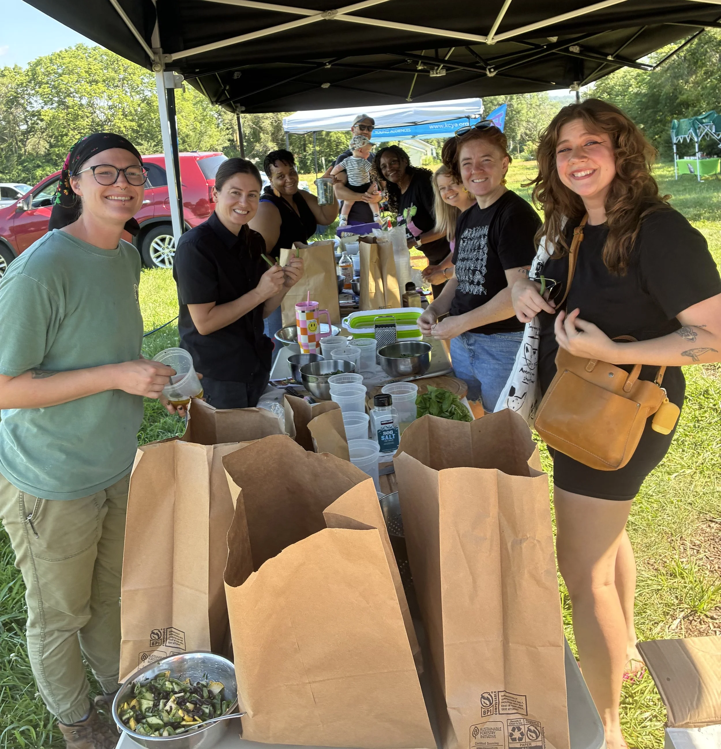 Group of women and a man smiling at an outdoor gathering under a black canopy, with a table of food, drinks, and paper bags, in a park-like setting with green trees and parked cars in the background.