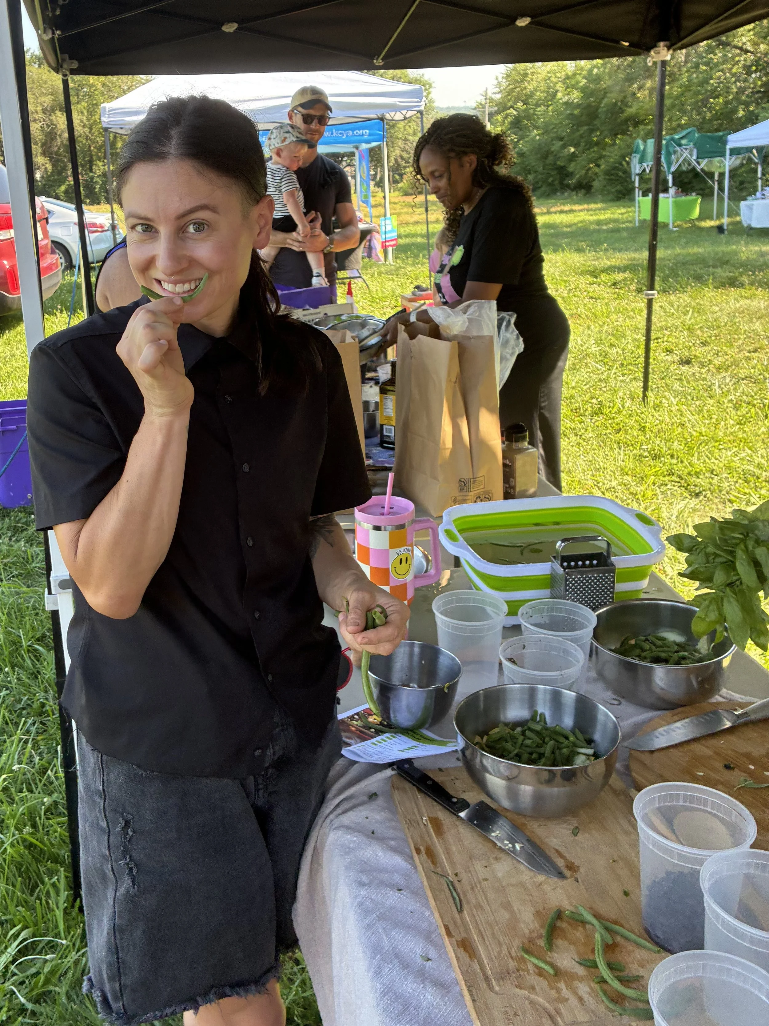 A woman smiling and tasting a green vegetable at a picnic table outside under a canopy. Other people are in the background preparing food, surrounded by green grass and trees.
