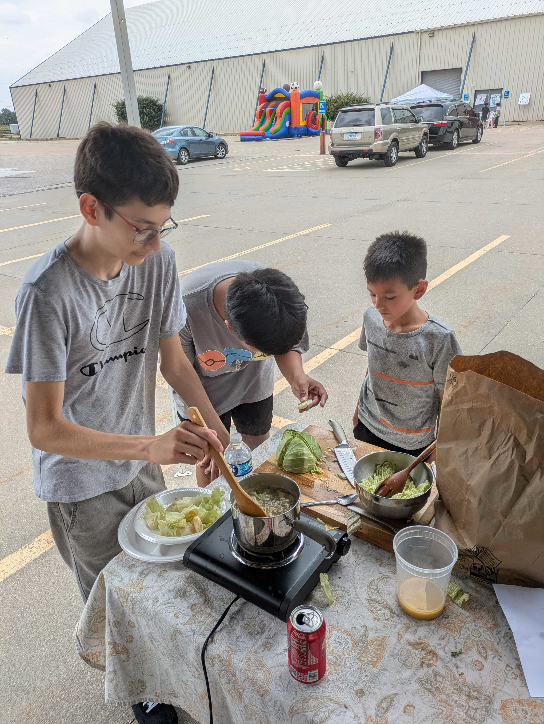 A group of children preparing food at an outdoor table with lettuce and a pot on a portable stove. A large paper bag and soda can are also on the table. In the background, there is a parking lot, several cars, a bounce house, and some people near a tent.