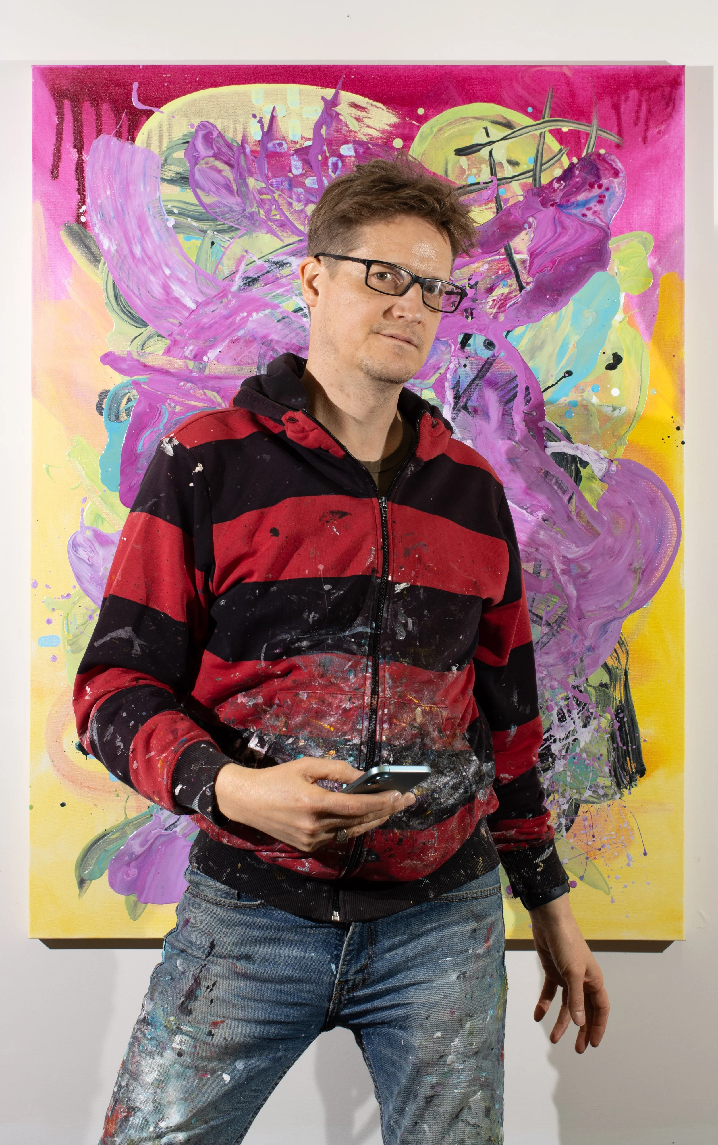 The painter Blake Brasher wearing a paint smeared red and black striped hoodie standing in front of a colorful abstract painting