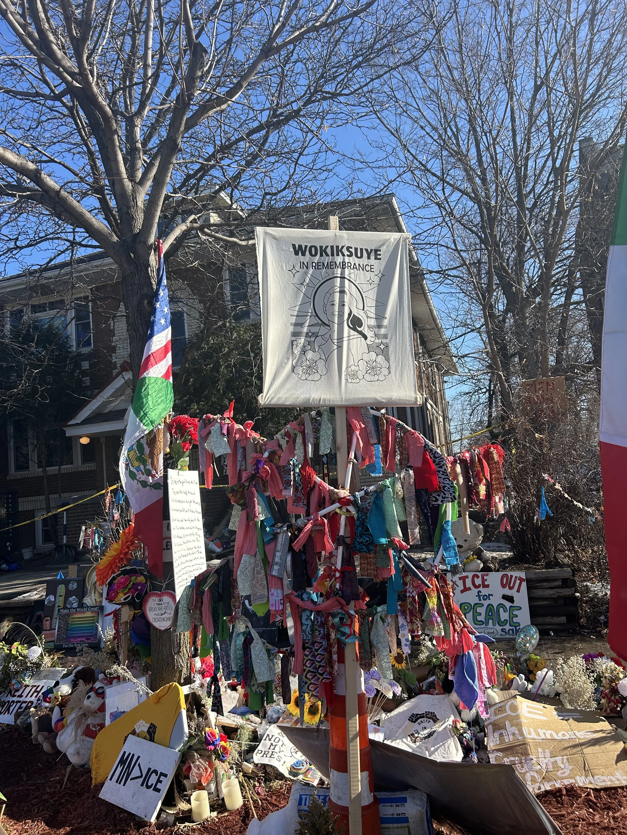 Image of a memorial outside with a sign saying "Wokiksuye" and then below the words "In Remembrance" next to an American Flag and multiple colorful prayer ties. On the ground are signs saying MN aginst ICE and flowers and stuffed animals.