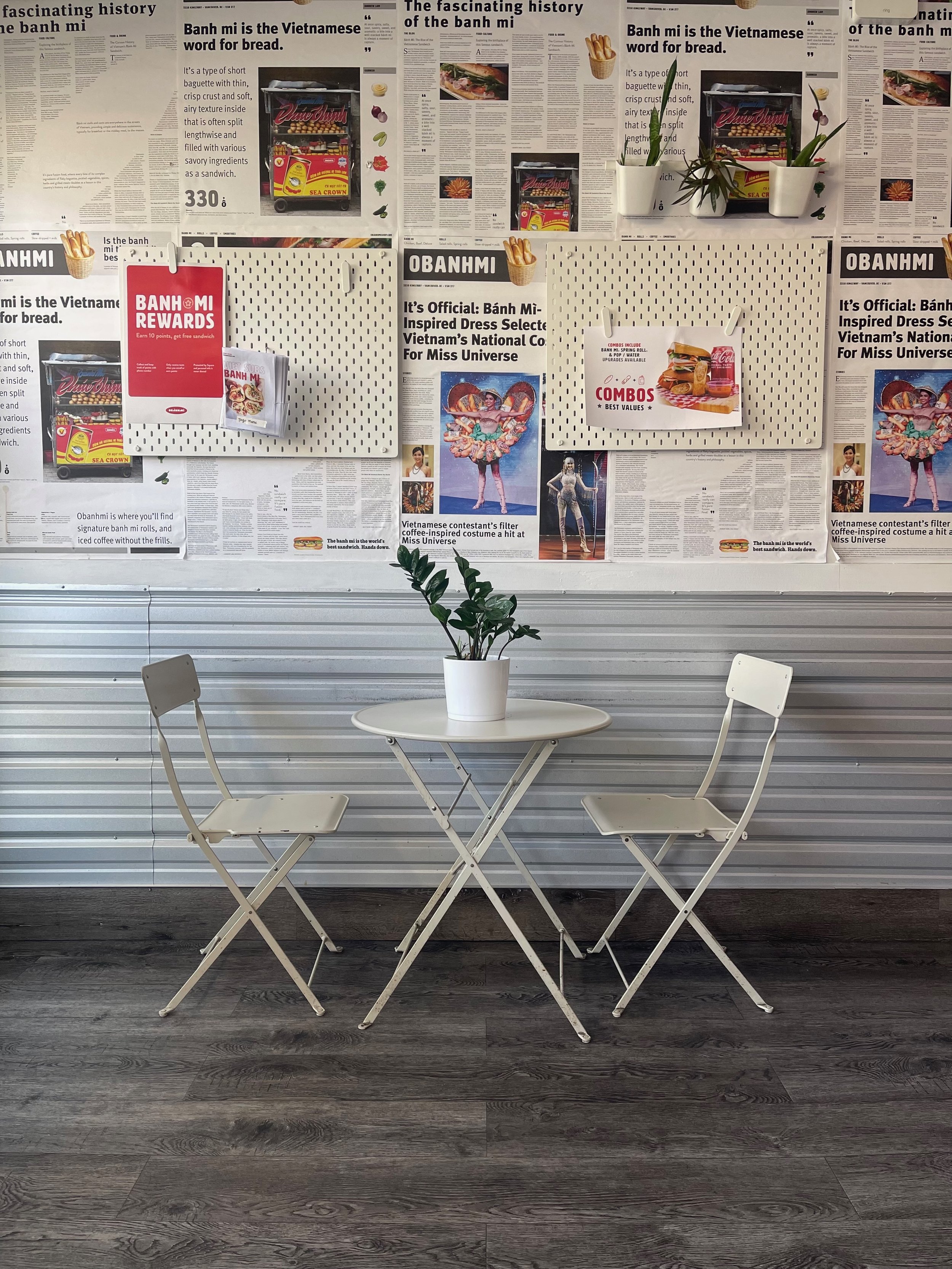A white round table with two white folding chairs in front of a wall decorated with newspaper clippings, potted plants, and promotional posters. This is Kingsway location.