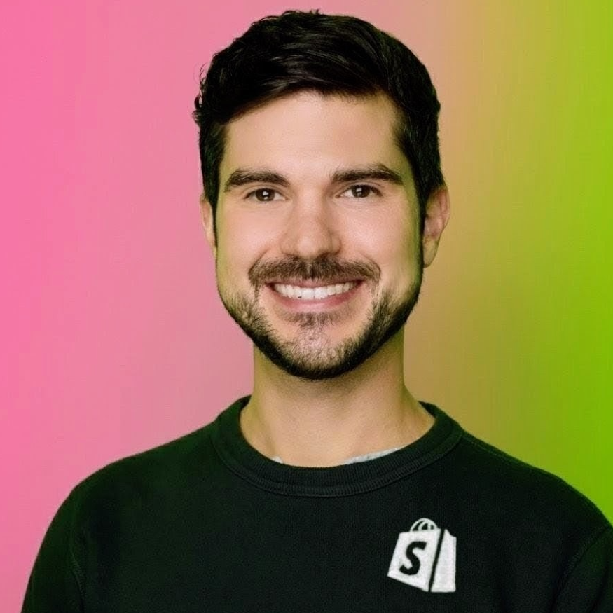 Justin D. Harris smiles facing the camera. He is wearing a black t-shirt with the Shopify logo on the chest.