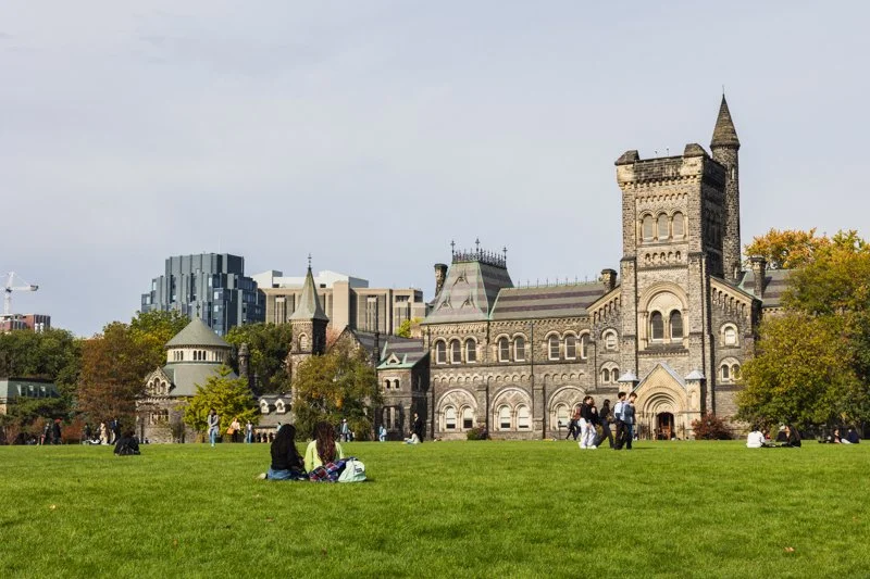 A wide view of U of T's historic stone building overlooking a large green lawn. Small groups of students sit and walk across the grass.