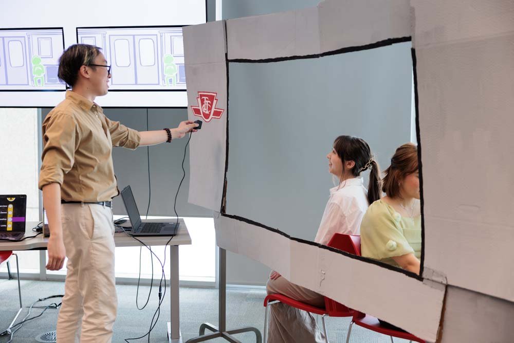 A student holds up a wired webcam in front of a mock TTC subway car made of cardboard. Two women are seated on the other side of a window opening in the cardboard.