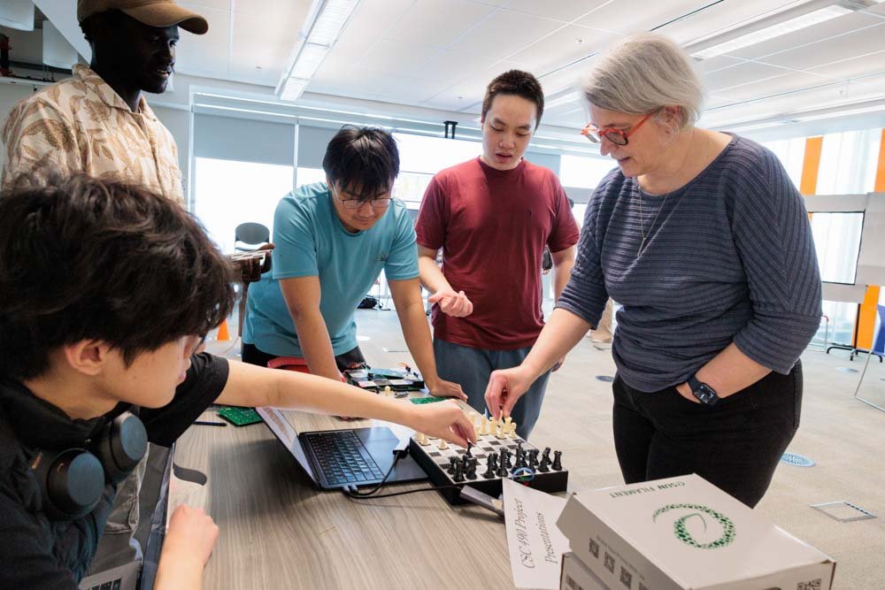 Five people around a chess board connected to a computer. Two of them are reaching for pieces on the chess board.
