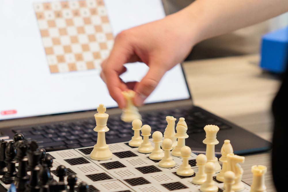 A hand moves a chess piece on a chess board, in front of a laptop screen that displays the position of the physical pieces on the board.