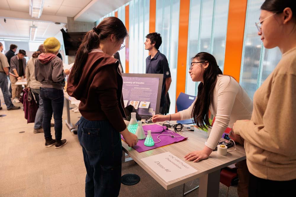 A student engages with a project involving plastic beakers with green LED lights, as two students look on.