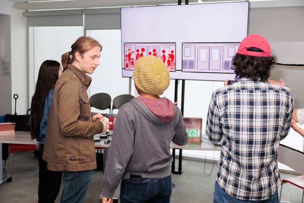 Three students from behind, with a TV screen in the background that has an illustration of full and empty subway cars.