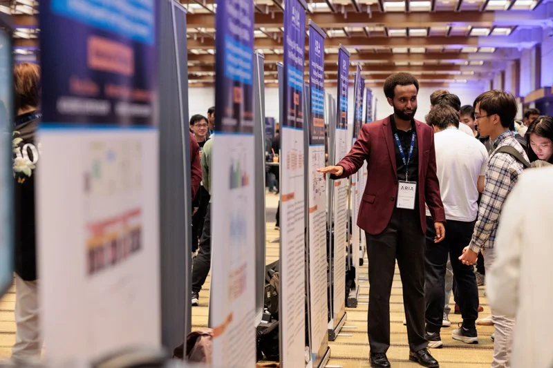  Person presenting a research poster in a large conference hall with multiple posters lined up and attendees walking through the exhibit area at the ARIA event. 