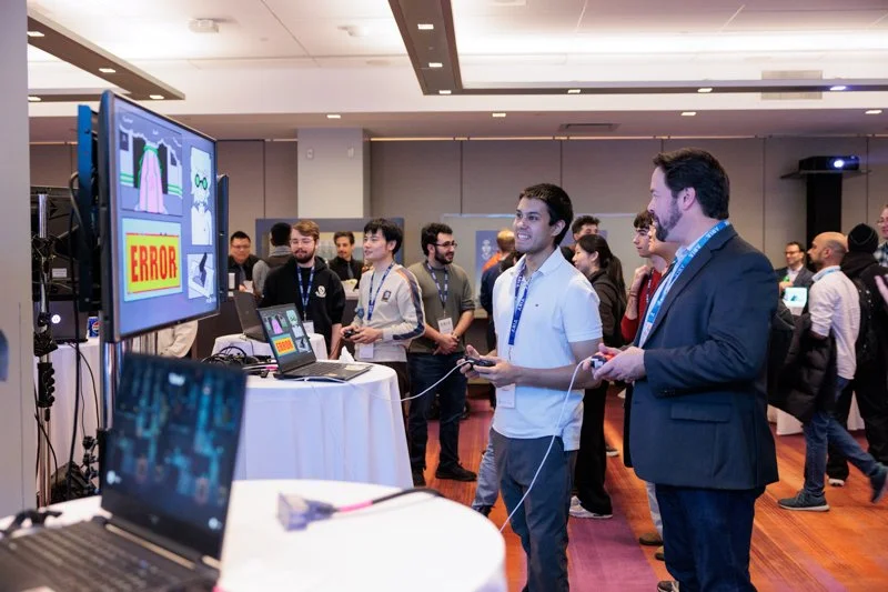  Two people using game controllers at an interactive station with a large screen displaying graphics and the word “ERROR,” while a group of attendees observes in the background at the ARIA event. 