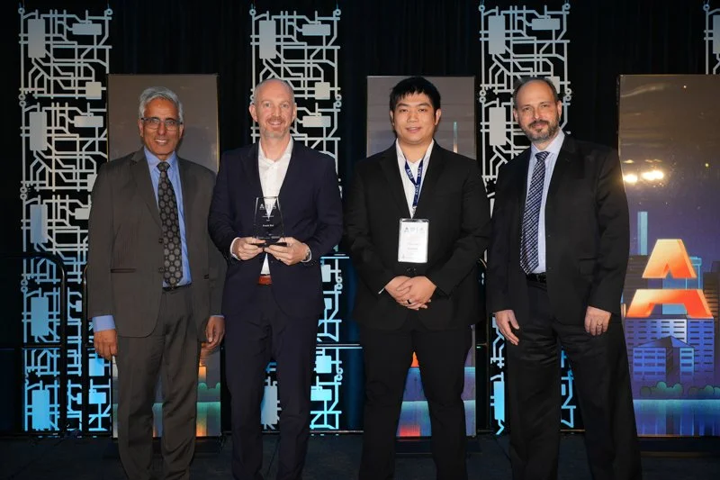  Four individuals in formal attire standing on a stage at the ARIA event, with one person holding an award. The backdrop features a circuit-themed design and illuminated panels.   