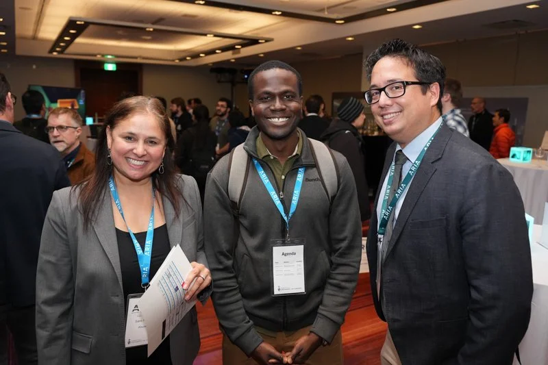  Three attendees wearing ARIA lanyards and badges standing together in a networking area at the event, with other participants and tables visible in the background. 