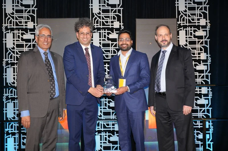  Four individuals in formal attire standing on a stage at the ARIA event, with two people holding an award together. The backdrop features a circuit-themed design and illuminated panels. 