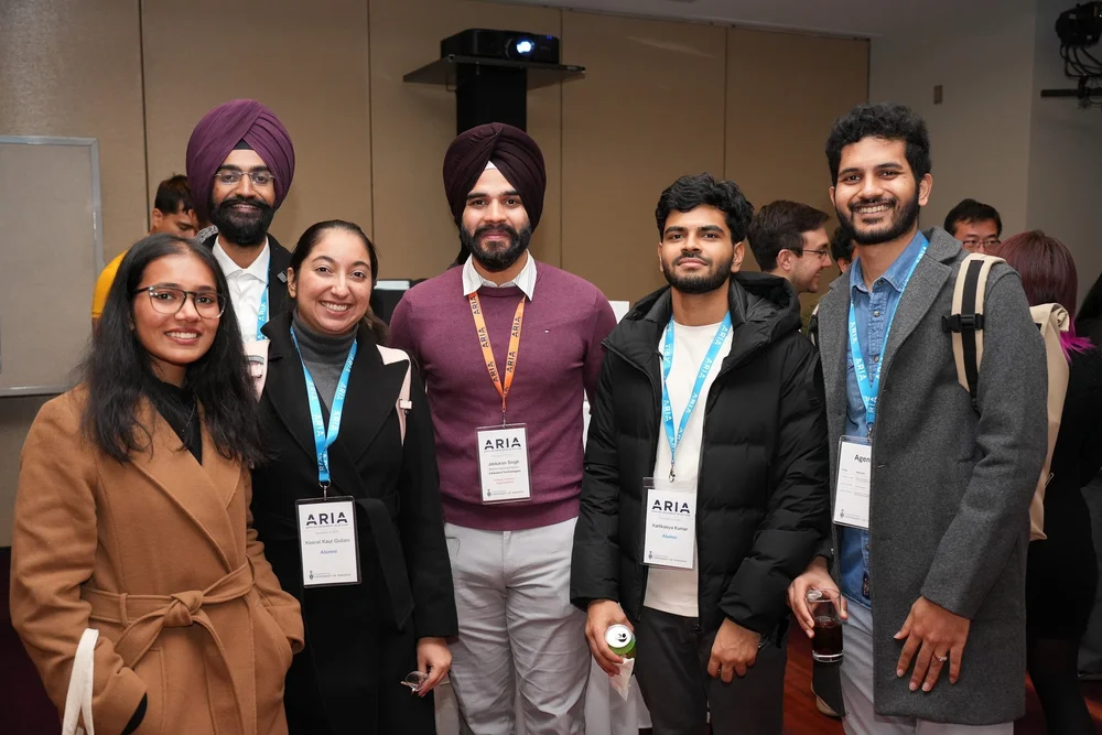  Group of attendees wearing ARIA lanyards and badges standing together in a networking area at the event, with others visible in the background. 