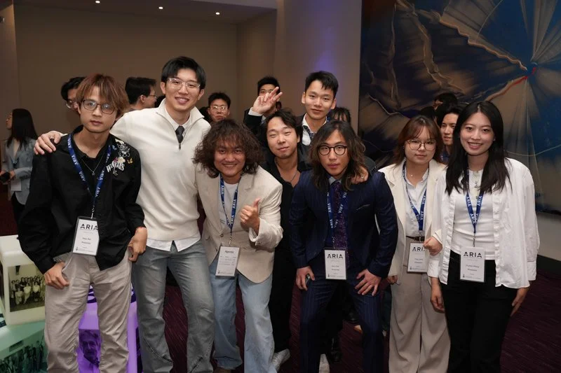  Group of attendees wearing ARIA lanyards and badges posing together in a networking area at the event, with colorful artwork visible on the wall in the background. 