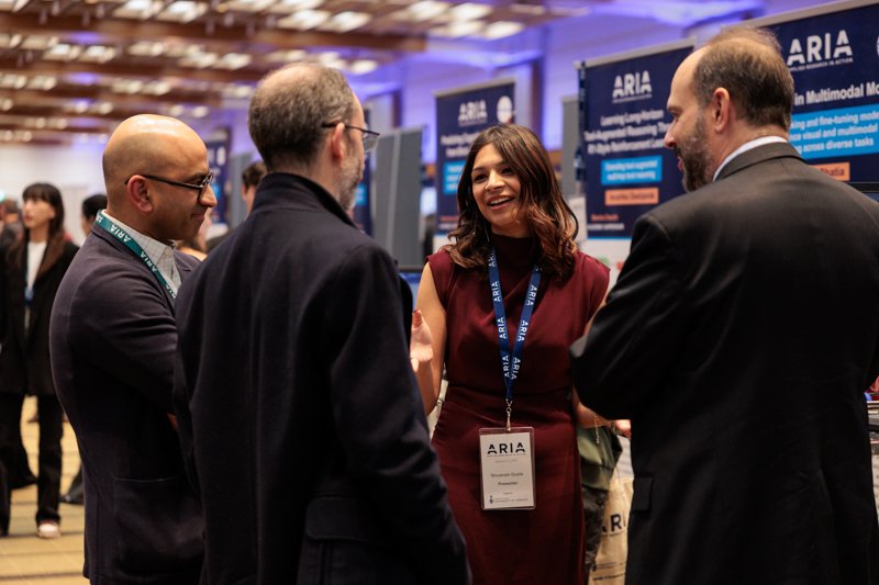  Group of attendees engaged in conversation in front of ARIA research posters at a conference exhibit area. 